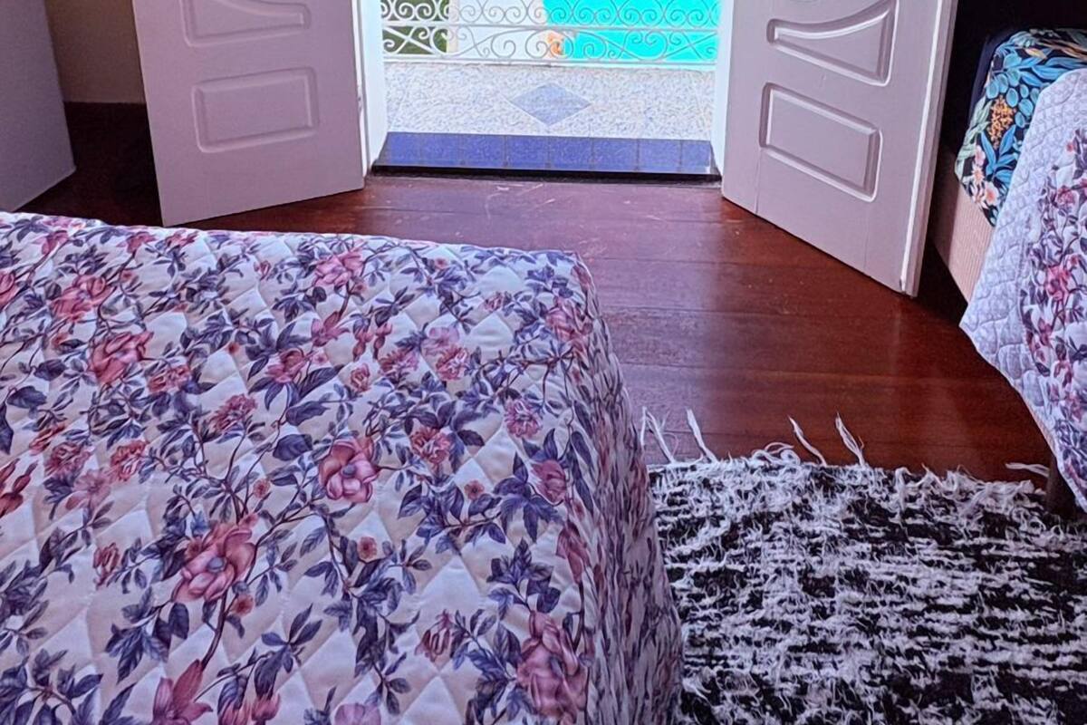 Une porte ouverte donnant sur un balcon avec une piscine, un lit recouvert d’une literie à motifs floraux et une vue sur un bâtiment verdoyant, surmonté d’un toit en tuiles.