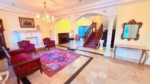 Un vaste salon doté d’une cheminée, de deux fauteuils, d’un piano et d’un escalier menant à l’étage supérieur.