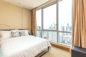 Ein Hotelzimmer mit einem großen Bett, einem Nachttisch, einer Lampe und Blick auf die Stadt durch große Fenster.