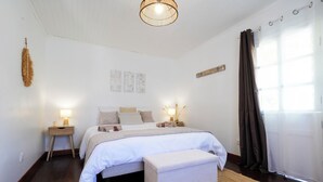 Un dormitorio con cama, mesitas de noche, lámpara colgante, ventana con cortinas y una obra de arte en la pared.