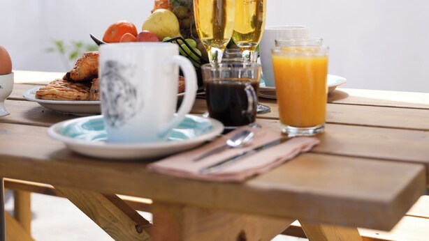 Una mesa con alimentos para el desayuno, como tostadas, fruta y bebidas, ubicada al aire libre con vistas a la vegetación y montañas.