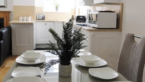 Una mesa de comedor dispuesta con platos y un centro de mesa con helecho, armarios blancos, un reloj y un microondas en una cocina luminosa.