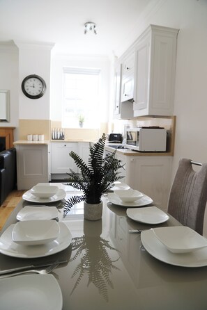 A dining table set with plates and a fern centerpiece, white cabinets, a clock, and a microwave in a bright kitchen. - Bright & Modern 2 Bed Apartment in the heart of Windsor - opposite the Castle (Windsor)