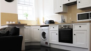Una cocina compacta con armarios blancos, lavadora, microondas y un sofá negro.