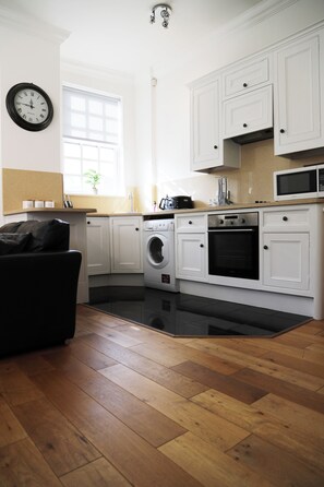 A compact kitchen with white cabinetry, a washing machine, a microwave, and a black sofa. - Bright & Modern 2 Bed Apartment in the heart of Windsor - opposite the Castle (Windsor)