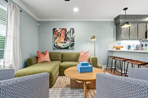 Ein Wohnzimmer mit Ecksofa, einem runden Couchtisch, einem Tigerbild, einer Küchenzeile mit Barhockern und einem Fenster mit Jalousien.