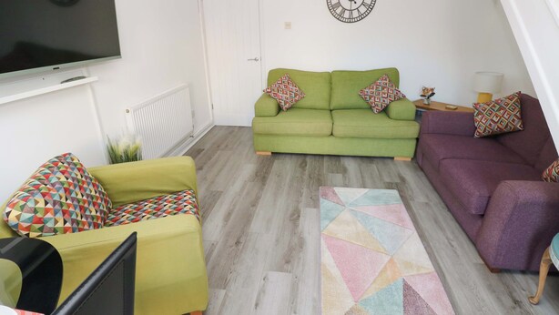 A living room with a green sofa, a purple sofa, a yellow armchair, a TV, and a clock on the wall. - The Gower Gateway (Swansea)