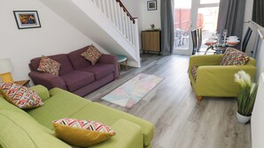 A living room with a staircase, green sofas, a dining area with a table set for two, and a view through a glass door. - The Gower Gateway (Swansea)
