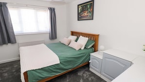A bedroom with a bed, a bedside table, a nightstand, a radiator, a window with curtains, and a painting on the wall. - The Gower Gateway (Swansea)