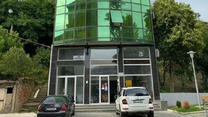 A modern building with a curved glass facade, a flat roof, and a parking area with two cars.