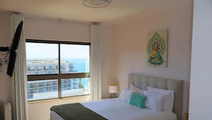 Ein Hotelzimmer mit einem großen Bett, einem Fenster mit Blick auf Gebäude, einem Gemälde an der Wand und einem an der Wand befestigten Fernseher.