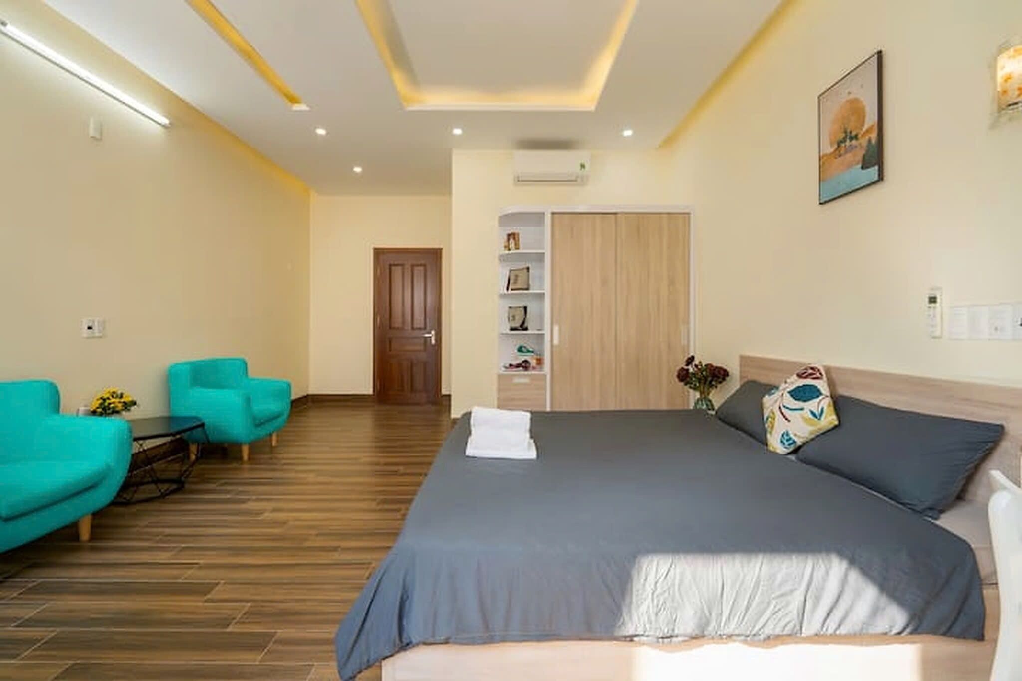 A hotel room with a large bed, a desk, two chairs, a wardrobe, and a painting on the wall.