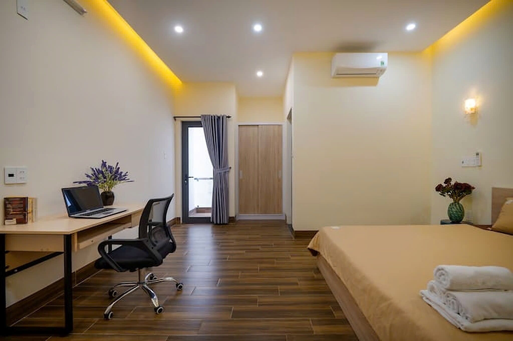 A hotel room with a wooden floor, a desk with a laptop, a bed with a beige bedspread, and a wall-mounted air conditioner.