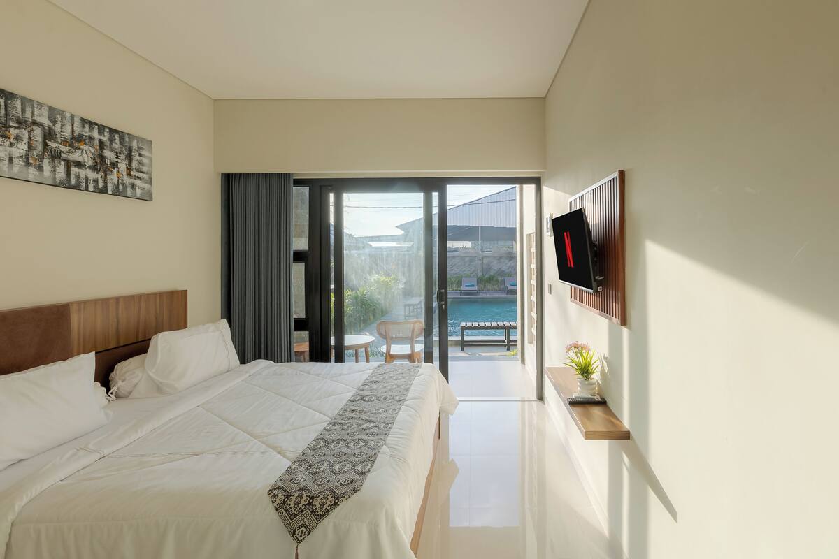 A hotel room with a bed, a TV, a small table with a plant, and a view of a pool and outdoor seating.