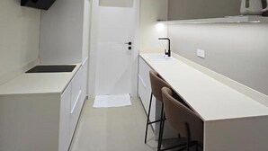 A compact kitchen with a white countertop, a black range hood, a white door, and a small bar stool. - Suites By Jordan's Hotel (Vlore)