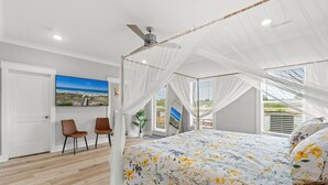 Ett sovrum med en himmelssäng, en takfläkt, ett stort fönster och en tavla med strandmotiv.