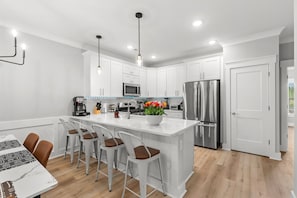 A modern kitchen with white cabinetry, stainless steel appliances, a central island with bar stools, pendant lighting, and a visible door to the right. - 5Kings waterfront Villa Near golf Beach Barefoot (North Myrtle Beach)