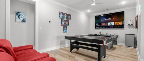 A modern living room with a pool table, red seating, a flat-screen TV displaying Google TV, and a ceiling fan.