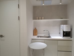 Una kitchenette compacta con un lavamanos, una cafetera, una mesita y una tabla de cortar de madera.