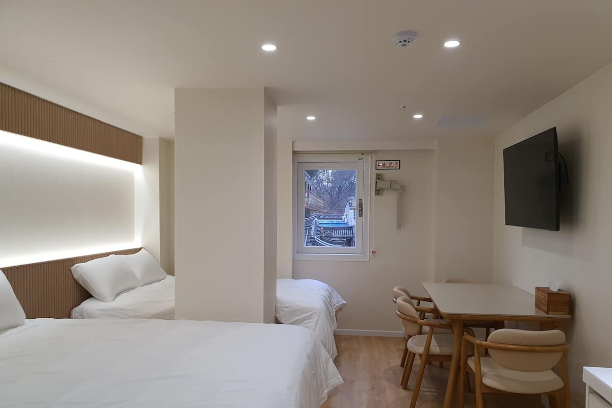 Una camera d'albergo con due letti, un tavolo da pranzo con sedie, una TV a parete e una finestra con vista sugli edifici.