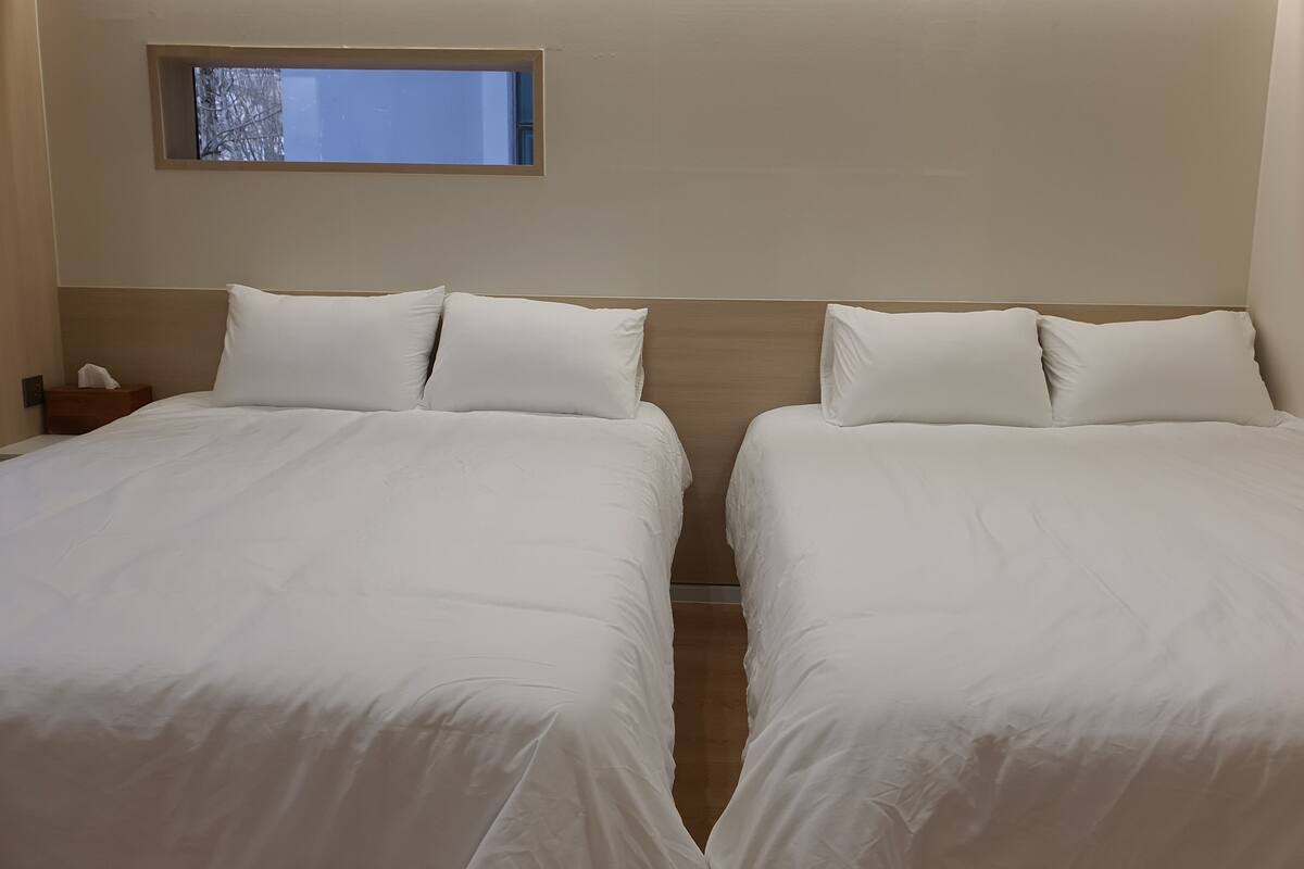 Camera d'albergo con due letti, un comodino, una piccola finestra e una testiera in legno.