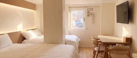 A hotel room with two beds, a desk with chairs, a television, and a window with a view of buildings.