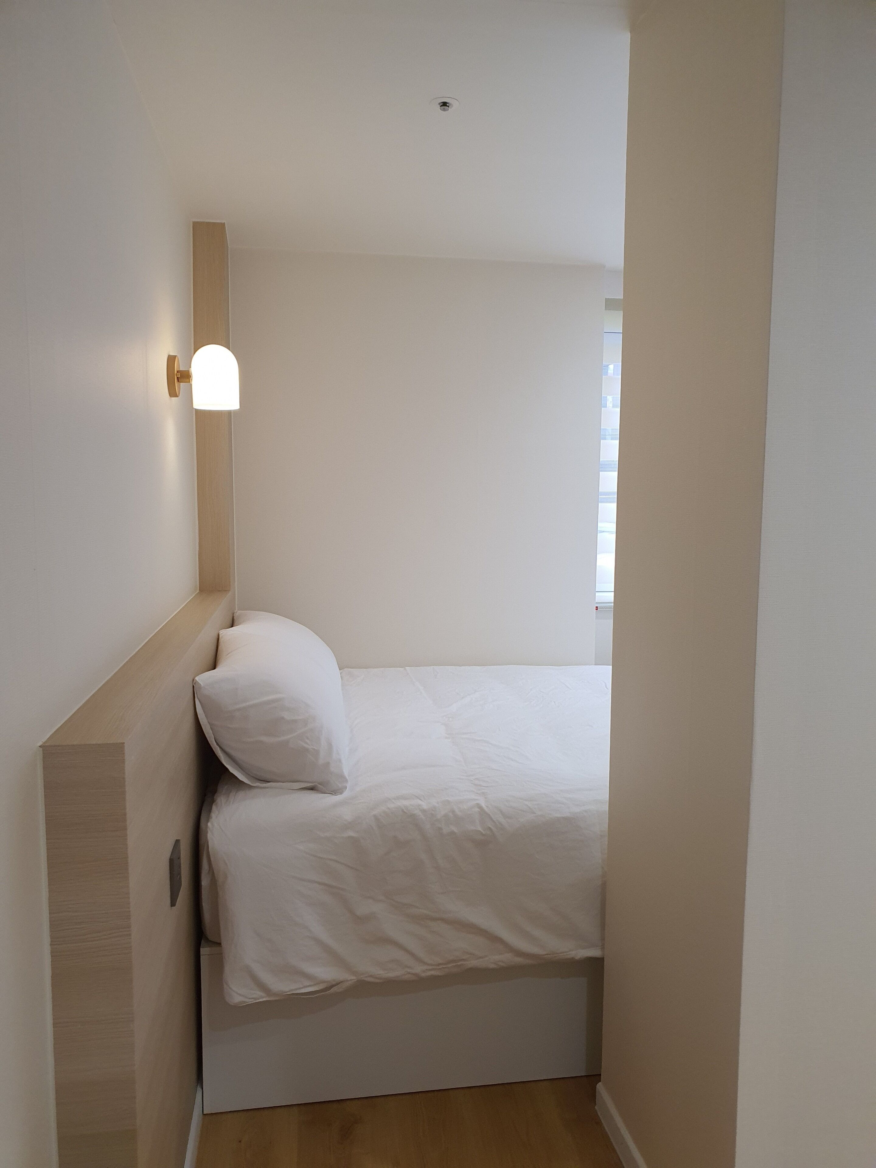 Ein kleiner, schmaler Raum mit einem Bett, einer Wandleuchte und einem Deckenlüfter.
