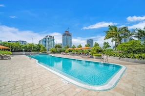 Une piscine dotée d’une terrasse carrelée, entourée de chaises longues et de parasols, avec la silhouette de la ville en arrière-plan.