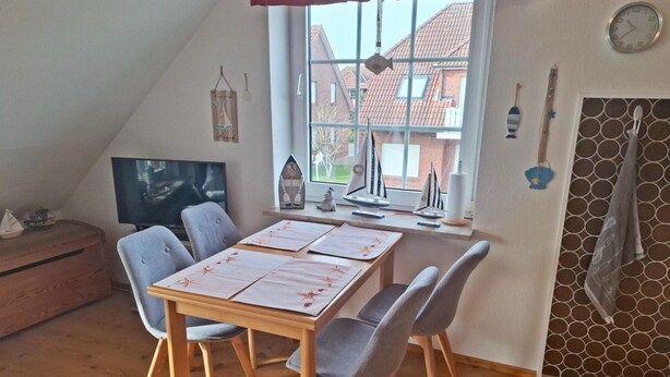 A dining area with a table, chairs, a TV, and a window with a view of buildings. - Ferienwohnung fur 4 Personen in Nessmersiel (Dornum)