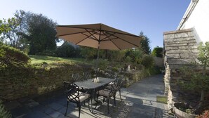 An outdoor patio with a table and chairs under a large umbrella, surrounded by stone walls and greenery. - Mays Cottage (Wadebridge)