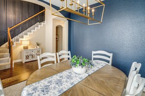 A dining area with a round table, white chairs, a floral table runner, a vase with green plants, and a staircase with wooden railings.