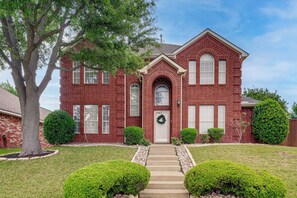 A red brick house with a front porch, a front door with a wreath, and a small front yard with trimmed bushes and a tree.