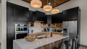 Una cucina moderna con mobili scuri, un'isola centrale, lampade a sospensione e un frigorifero in acciaio inossidabile.