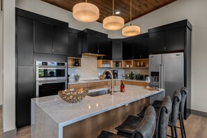 A modern kitchen with dark cabinets, a central island, pendant lights, and a stainless steel refrigerator. - Summit Splendor | Steps to The Trillium, Indoor Pool, Playground, Views! (Sevierville)