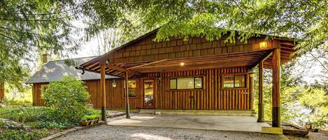 A wooden cabin with a porch, surrounded by trees and a gravel driveway.