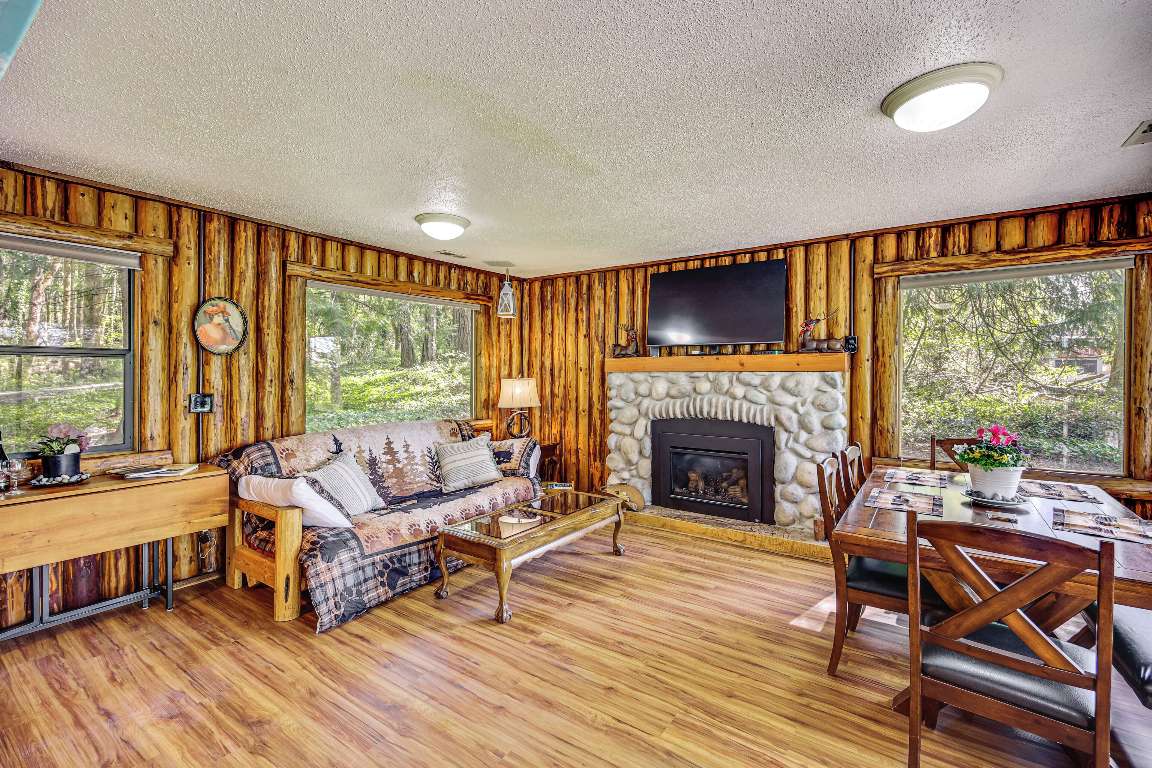 A rustic cabin interior with a stone fireplace, wooden walls, a sofa, a dining table, and large windows.