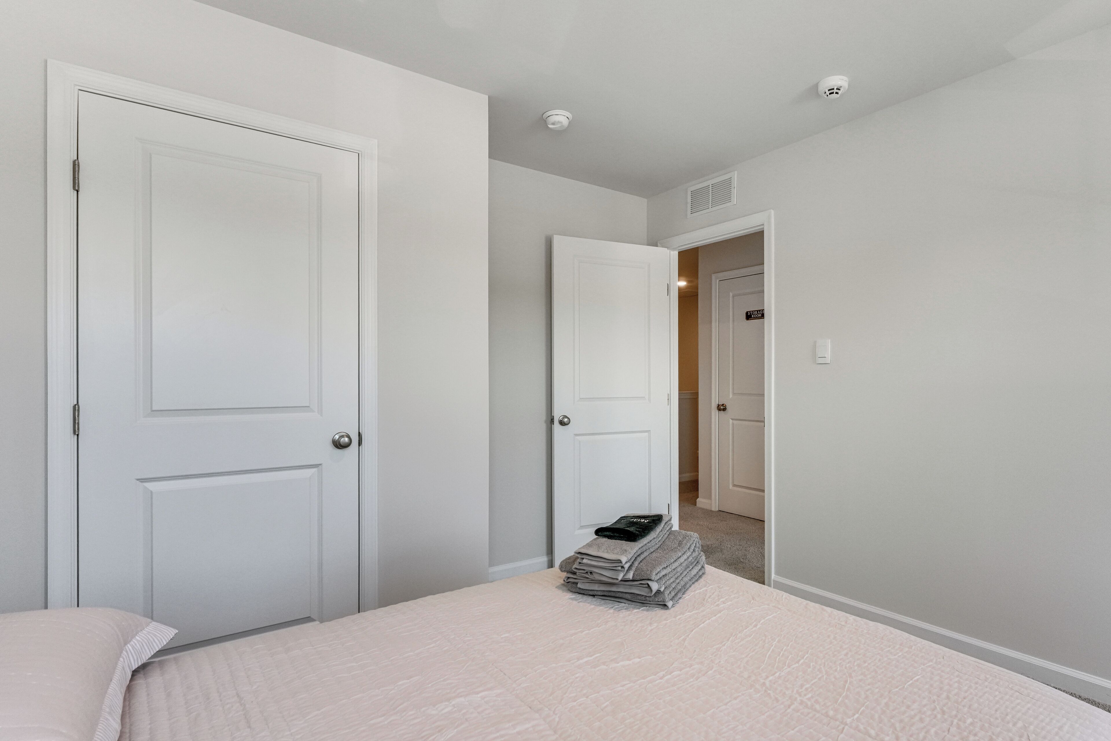 Een slaapkamer met twee witte deuren, een bed met een lichtgekleurd dekbed en een stapje opgevouwen handdoeken op het bed.