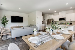 A modern living room with a dining area set for a meal, a flat-screen TV, and a kitchen with white cabinets and appliances.