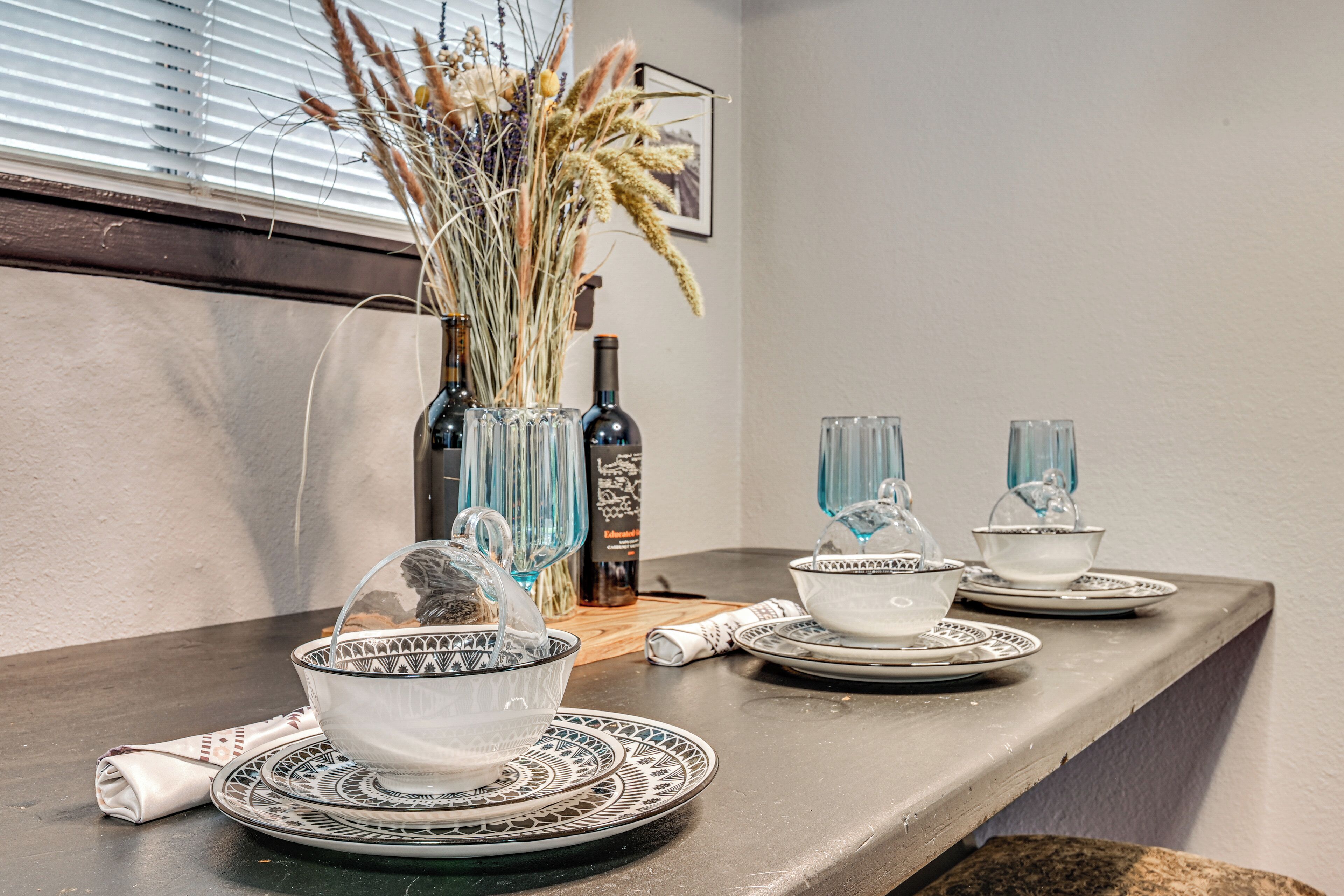 A dining table with a vase of dried plants, a bottle, and glassware.