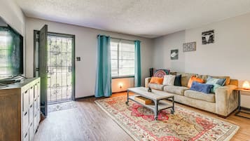 A living room with a sofa, coffee table, and a TV. There's a door leading outside, a window with curtains, and a rug on the floor.
