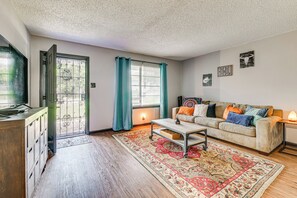 Ein Wohnzimmer mit einer Couch, einem Couchtisch und einem Fernseher. Eine Tür führt nach draußen, ein Fenster mit Vorhängen und ein Teppich liegen auf dem Boden.