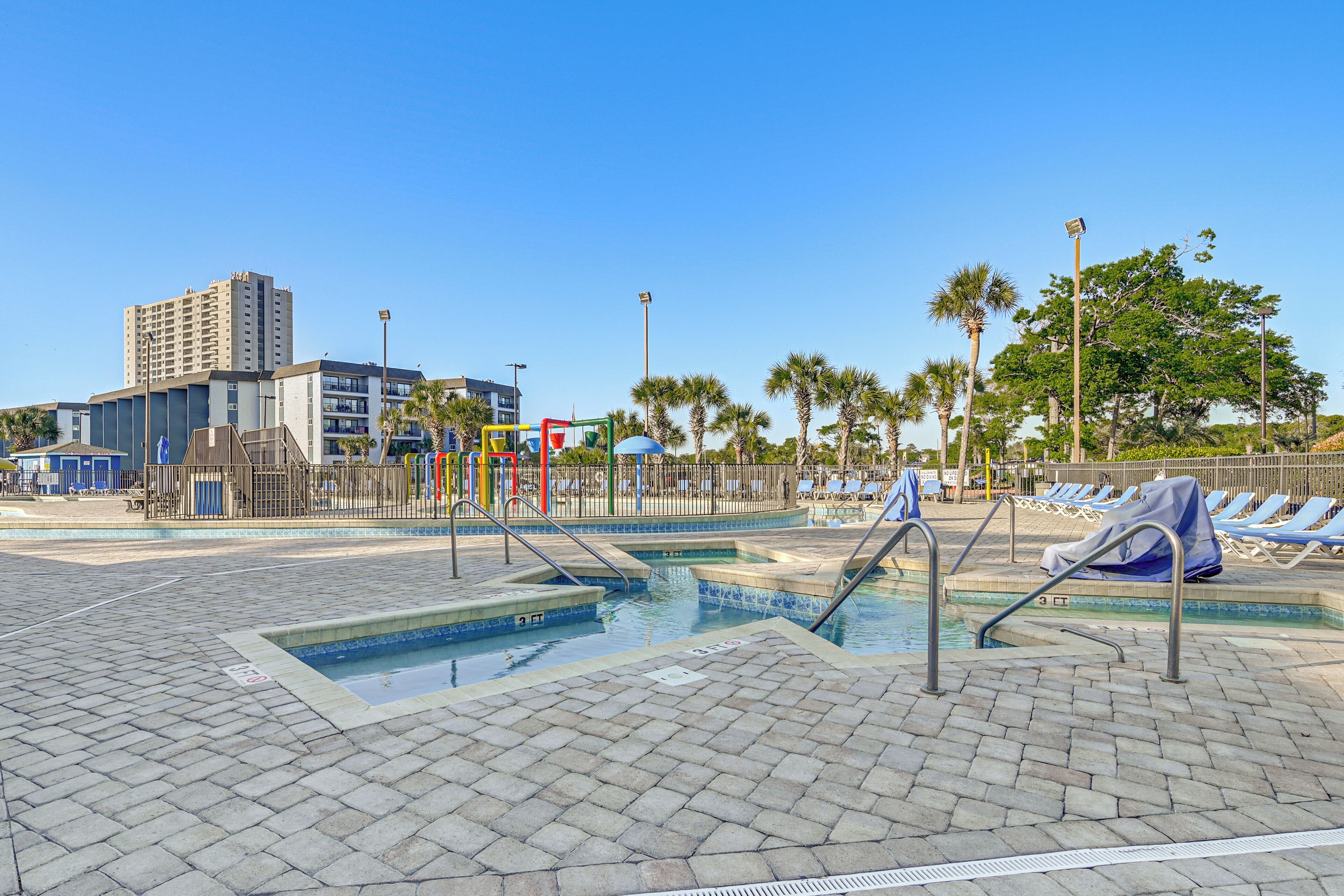 A swimming pool area with a children's play structure, lounge chairs, and a tall building in the background.