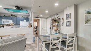 Een moderne keuken met een magnetron, kasten en een bordje met de tekst 'Keuken'. Er is een eethoek met een tafel en stoelen, en een gang die leidt naar een kamer in strandthema.