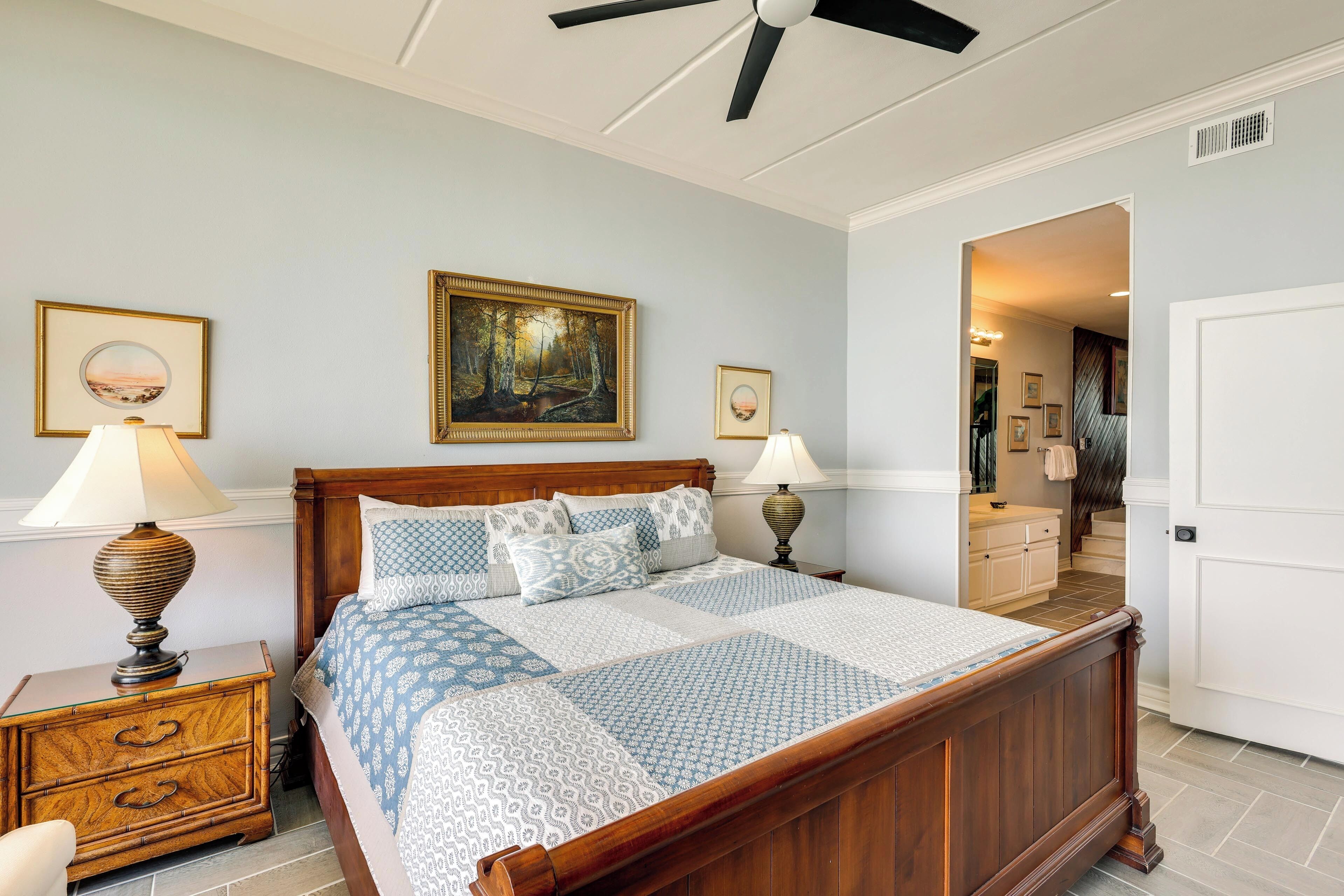 A bedroom with a wooden bed, two bedside lamps, a ceiling fan, and a framed painting on the wall.