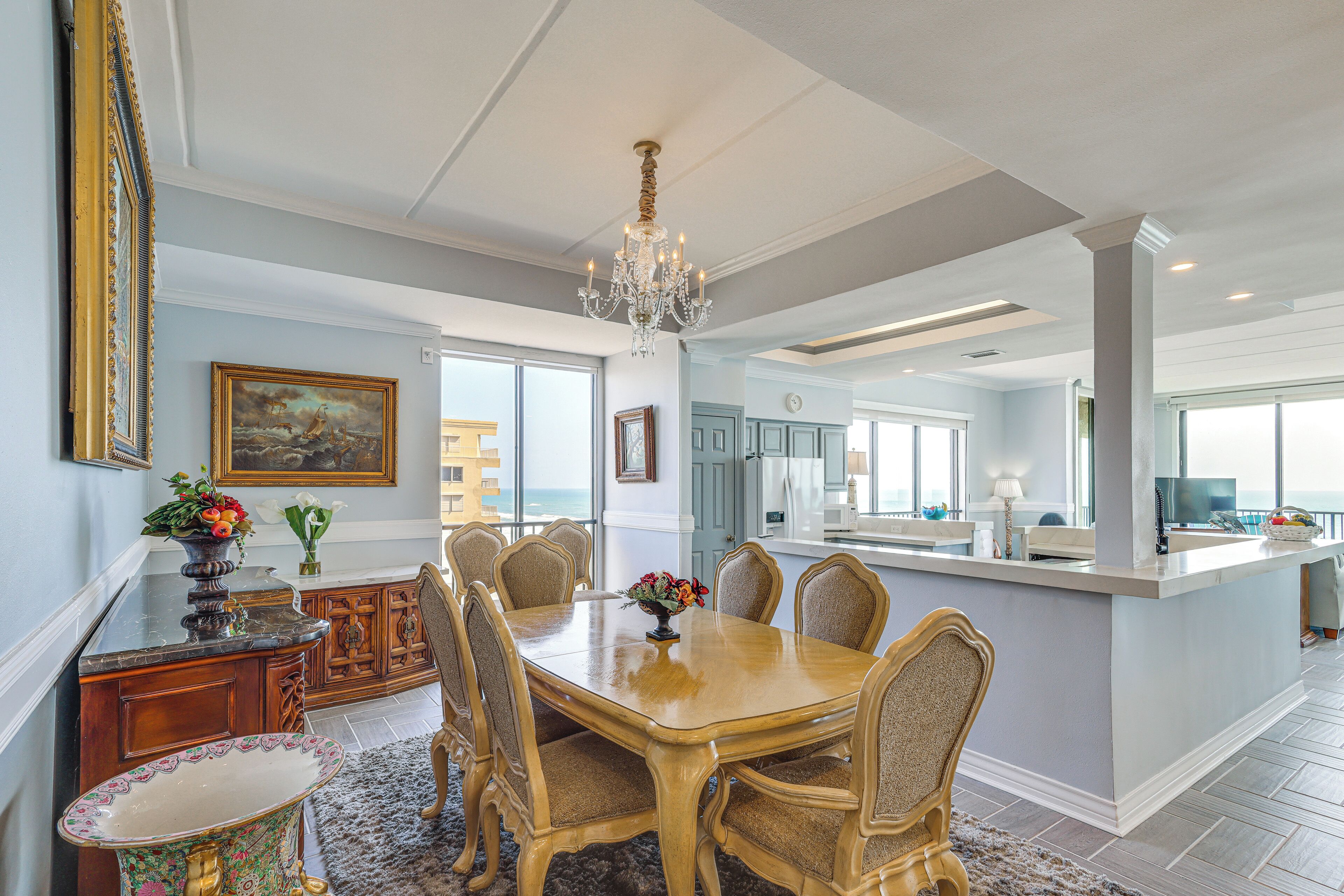 A dining area with a large wooden table, chairs, a chandelier, and a view of the ocean.