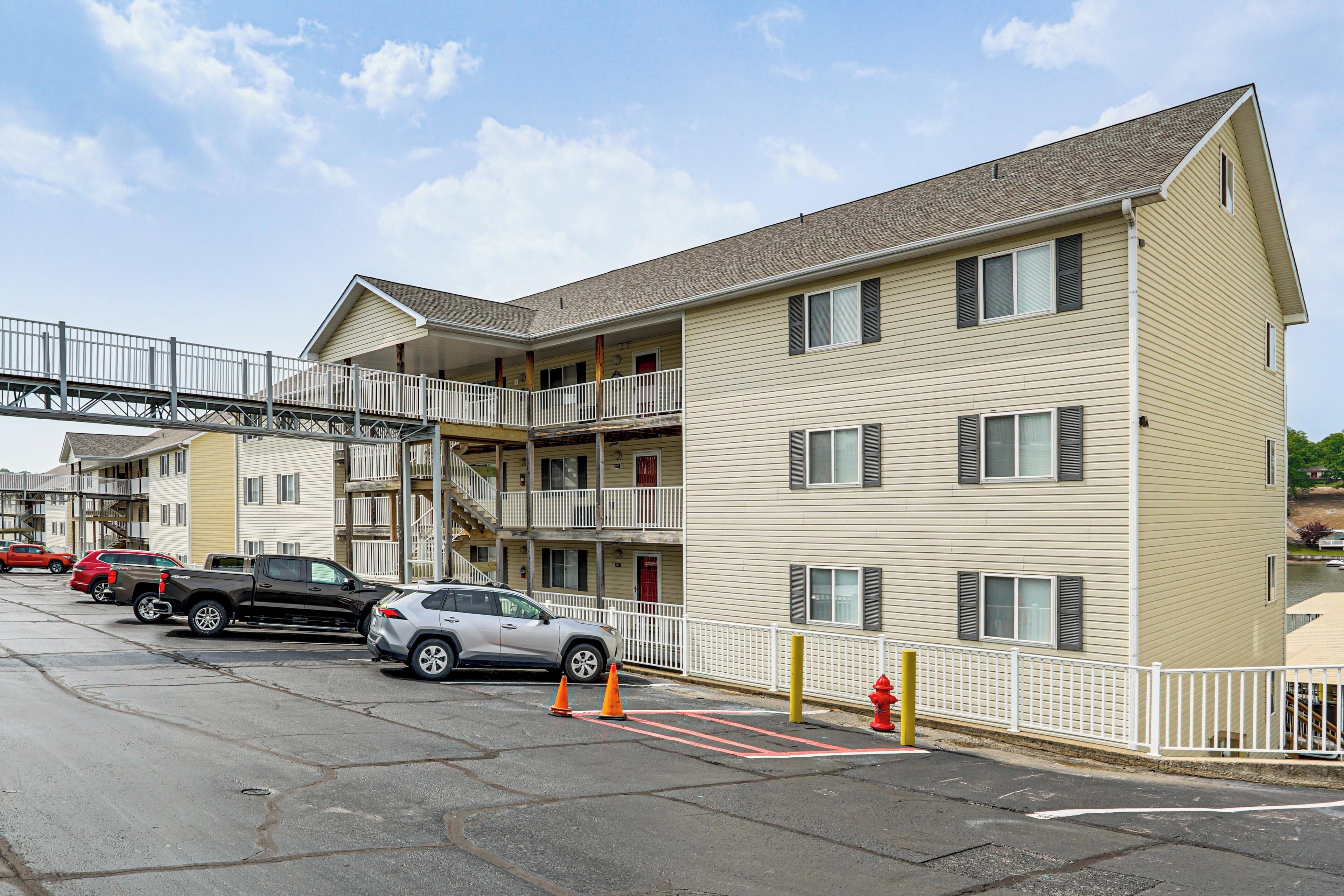 A multi-story building with a balcony, a parking lot with cars, and a pedestrian bridge.