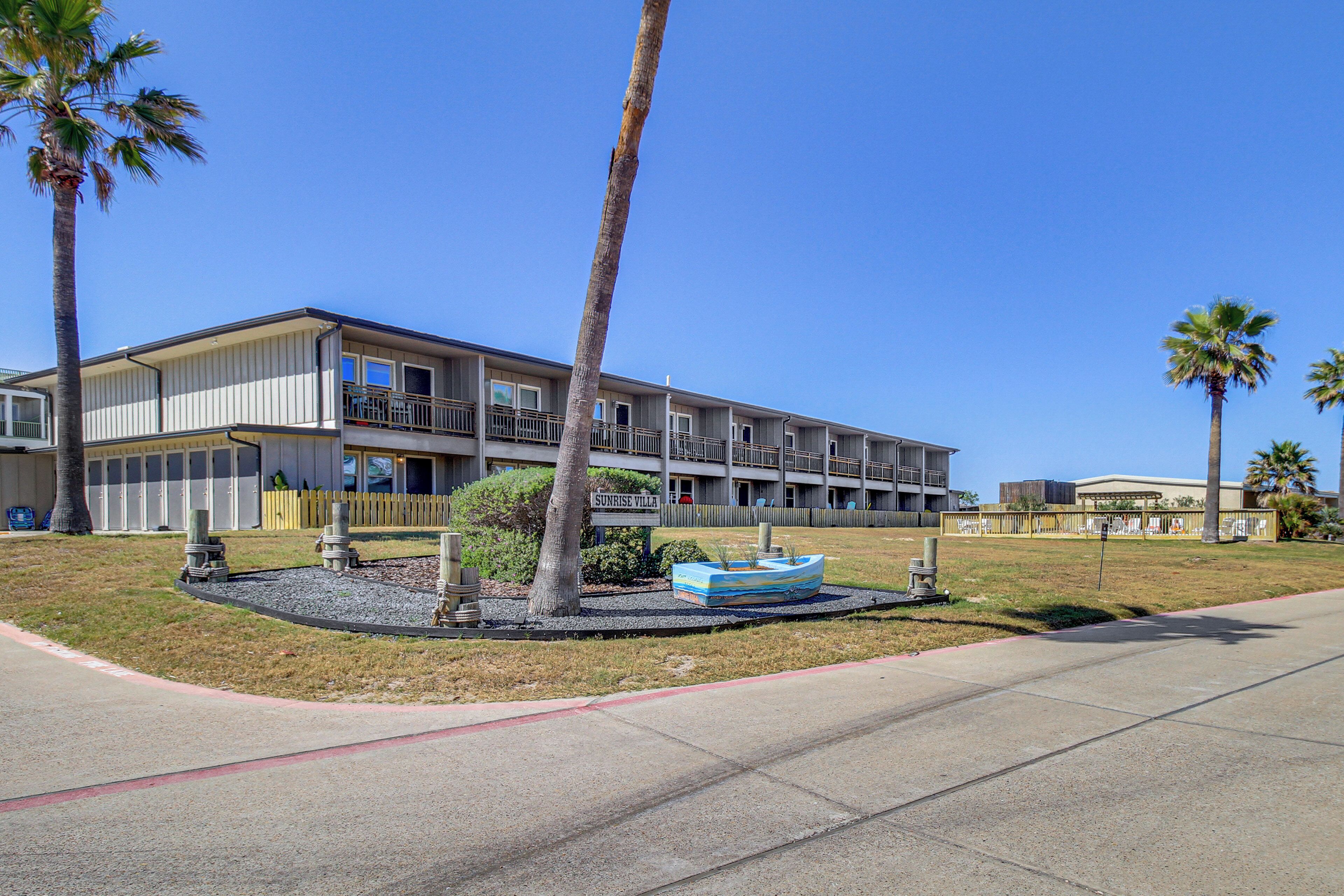 A multi-story hotel with a flat roof, balconies, and a landscaped area with palm trees.