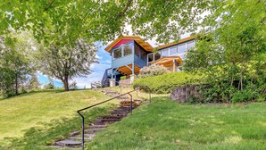A modern house with a staircase leading to a wooden deck, surrounded by lush greenery. - Stunning Sandpoint Lakefront Home: Hot Tub + Dock (Sandpoint)