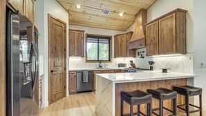 A modern kitchen with wooden cabinets, a stainless steel refrigerator, a marble island with bar stools, and a window providing natural light. - Modern Luxury in Torreon w/ HOT Tub! (Show Low)