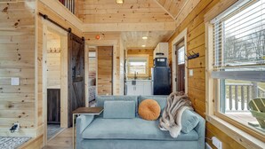 A cozy cabin interior with a sofa, a table, and a window overlooking a deck. - Handcrafted Tiny Home, Near Dtwn Boone (Boone)